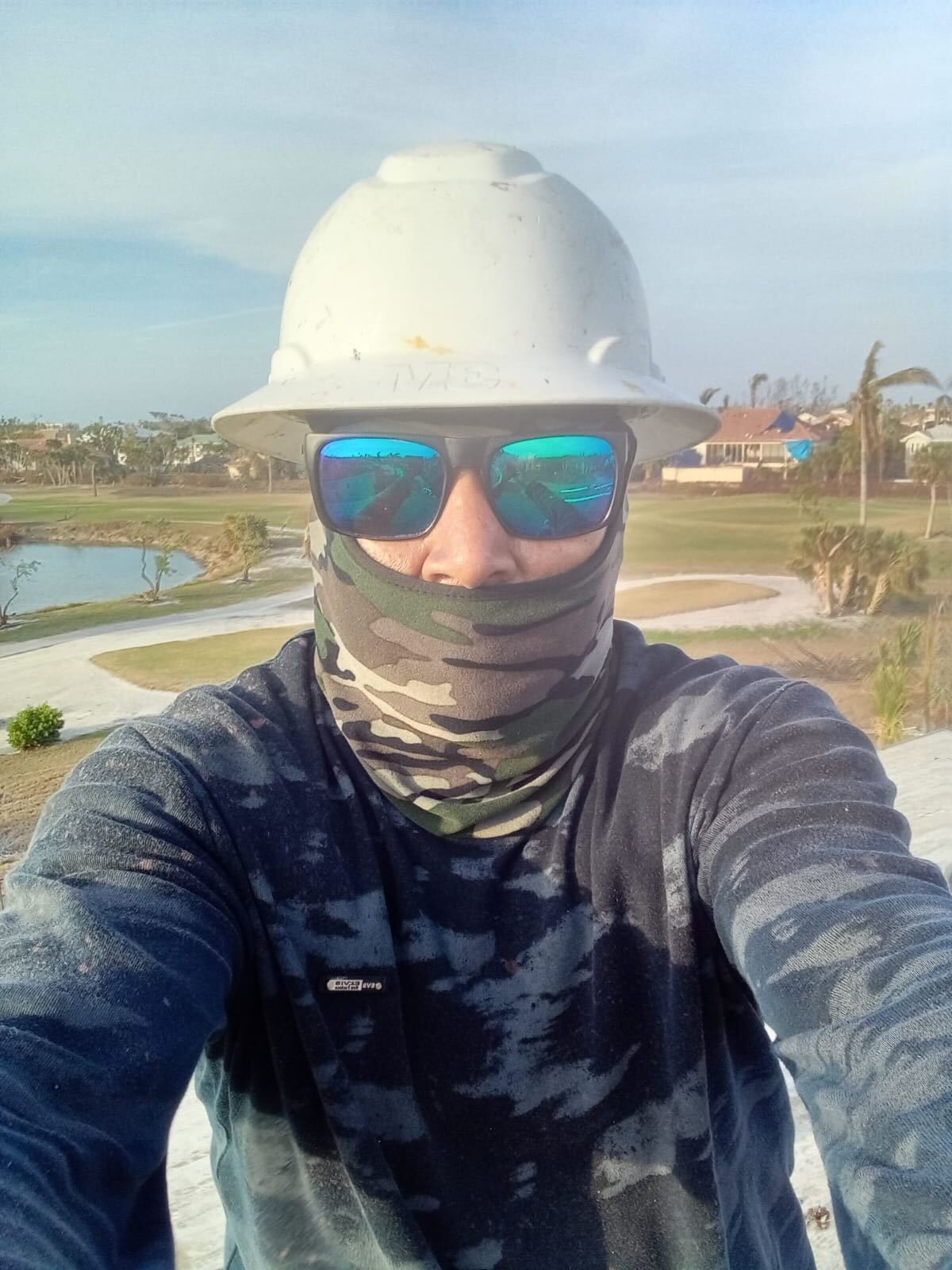 Marcos, trabajador de restauración después de desastres climáticos como huracanes, inundaciones y fuegos forestales, se toma una foto en su trabajo en Fort Myers, Florida. Marcos tenía puesto un casco de seguridad color blanco, gafas de sol y una cobertura de cara de camuflaje y una camisa con el mismo estampado. (Cortesía de Marcos)